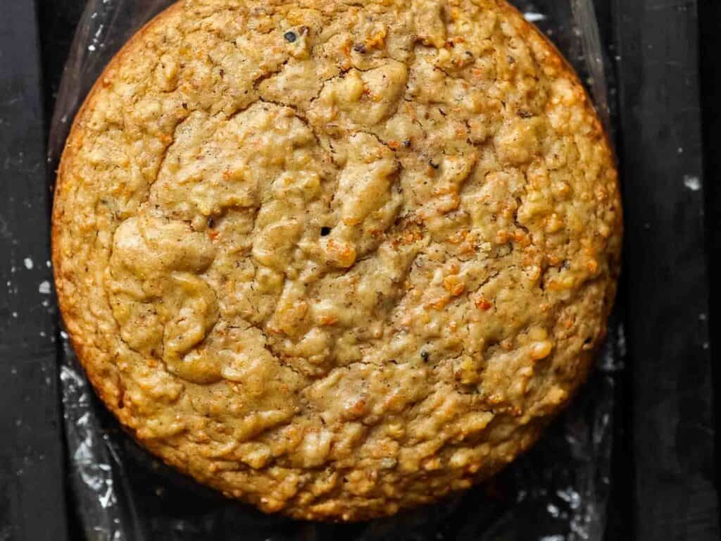 A close-up of a round, golden-brown vegan carrot cake with a textured surface, resting on a dark background.